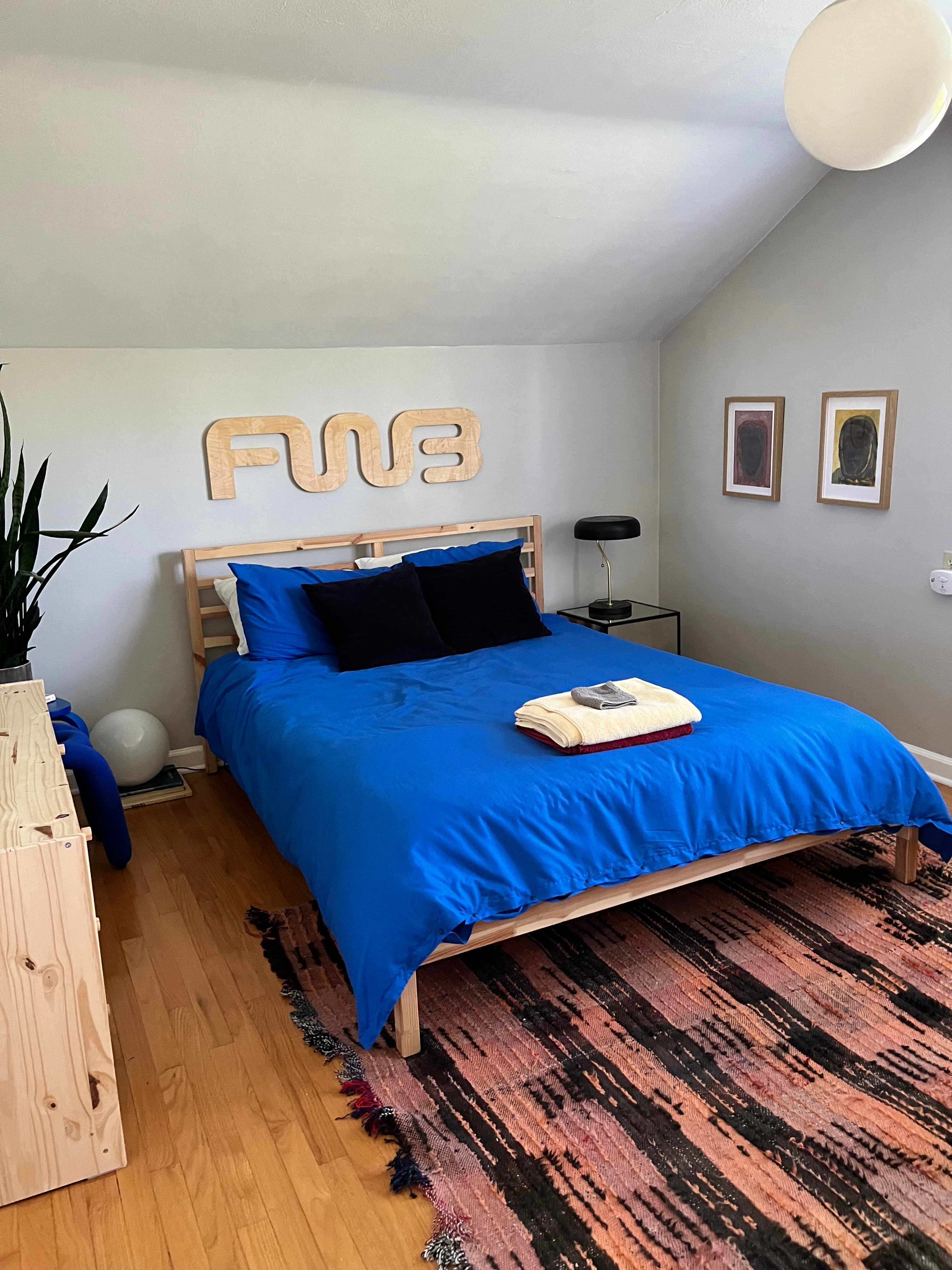 Minimalist bedroom with a blue bed, wooden FWB sign, and a pink patterned rug.