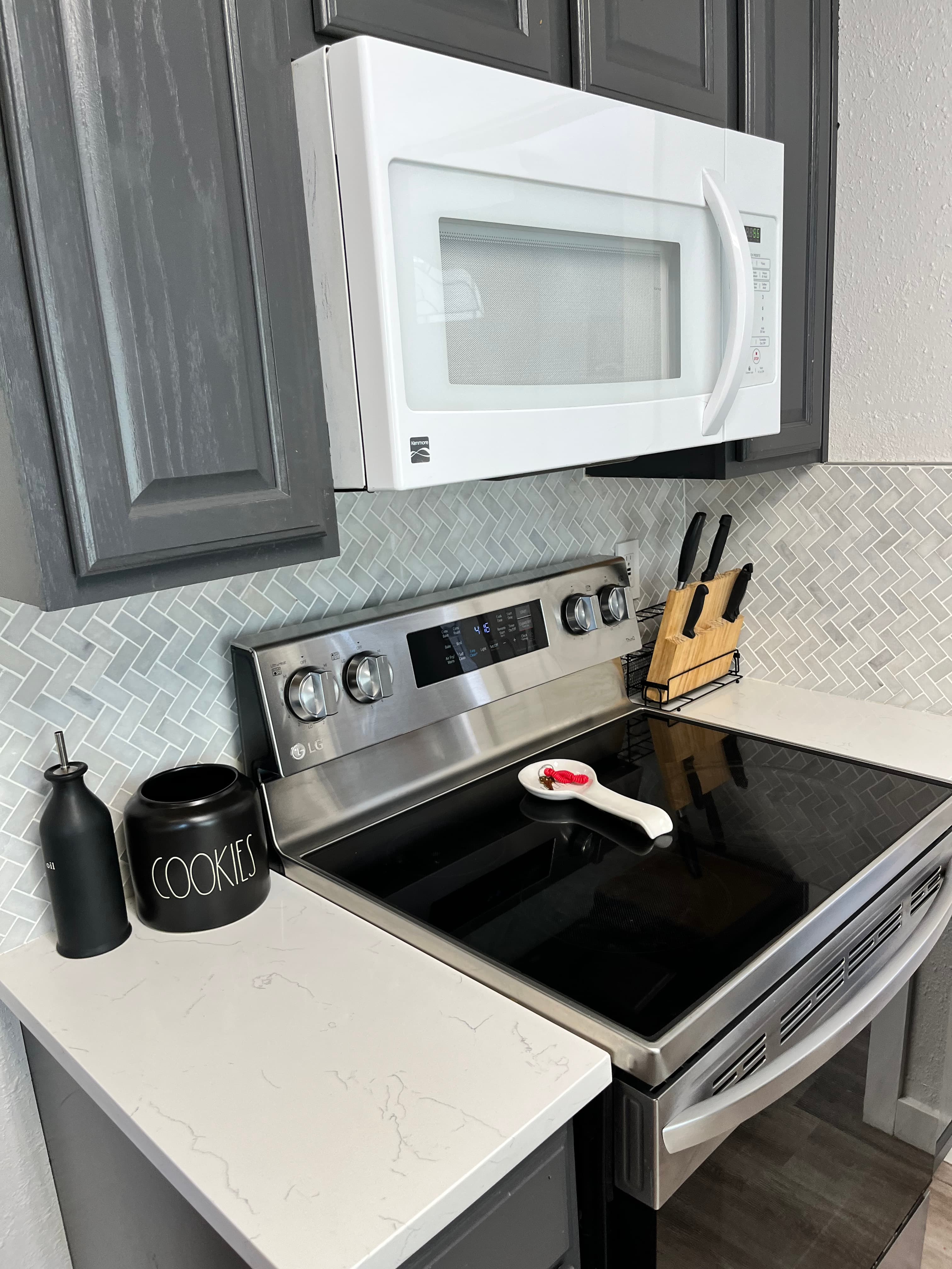 Stainless steel stove and white microwave with dark grey cabinets and herringbone tile backsplash.